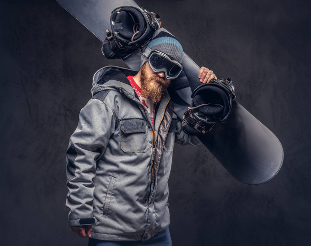 Brutale roodharige snowboarder met een volle baard in een winter-hoed en beschermende bril gekleed in een snowboard jas houdt een snowboard op zijn schouder in een studio. Geïsoleerd op een grijze achtergrond. - Foto, afbeelding