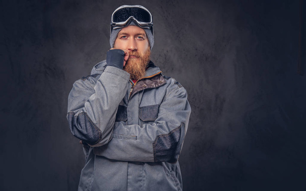 Ritratto di una rossa pensosa snowboarder con la barba piena in un cappello invernale e occhiali protettivi vestiti con un cappotto da snowboard in posa in uno studio, distogliendo lo sguardo. Isolato su uno sfondo scuro strutturato
. - Foto, immagini