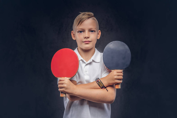 Cute boy dressed in a white t-shirt holds two ping-pong racquets in crossed hands. Isolated on a dark textured background. - Fotoğraf, Görsel