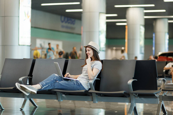 Young pensive traveler tourist woman in hat working on laptop while waiting in lobby hall at international airport. Passenger traveling abroad on weekends getaway. Air travel, flight journey concept - Photo, Image