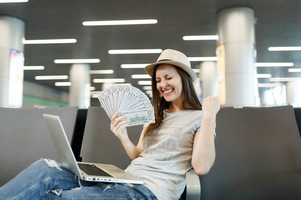Young happy traveler tourist woman with laptop hold bundle of dollars cash money, doing winner gesture wait in lobby hall at airport. Passenger traveling abroad on weekend getaway. Air flight concept - Foto, Imagem