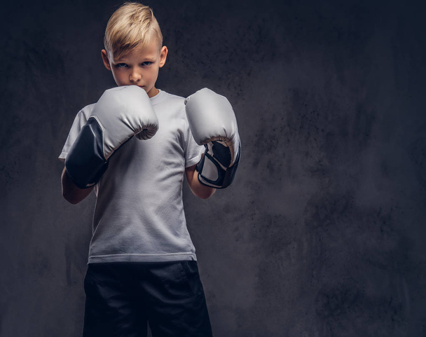 Un guapo boxeador de niño con el pelo rubio vestido con una camiseta blanca con guantes listos para pelear. Aislado sobre el fondo de textura oscura
. - Foto, imagen