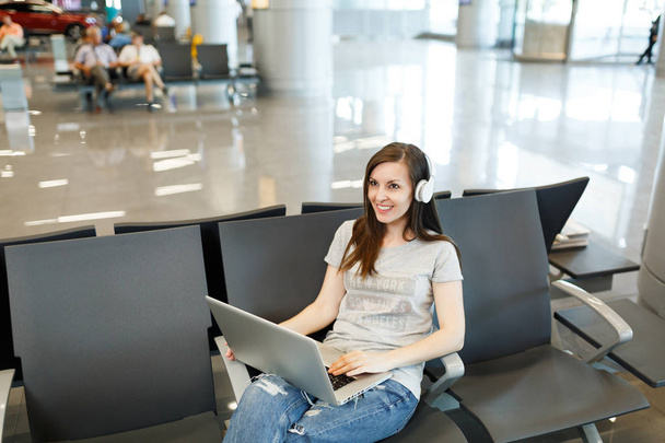 Young smiling traveler tourist woman with headphones listening music working on laptop, wait in lobby hall at international airport. Passenger traveling abroad on weekends getaway. Air flight concept - Photo, Image