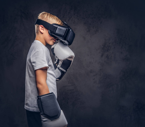 Schoolboy boxer with blonde hair dressed in a white t-shirt wearing visual reality glasses and boxing gloves, workout in a studio. Isolated on a dark textured background. - Foto, immagini