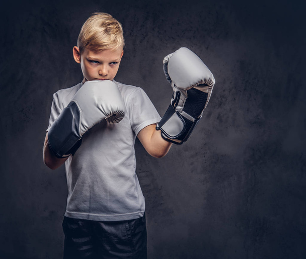 ein hübscher kleiner Boxer mit blonden Haaren, bekleidet mit einem weißen T-Shirt und Handschuhen, bereit zum Kampf. isoliert auf dem dunklen strukturierten Hintergrund. - Foto, Bild