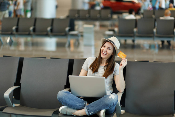 Молодая улыбчивая туристка-путешественница в шляпе сидит со скрещенными ногами, работая на ноутбуке, держит кредитную карту в холле аэропорта. Пассажир, путешествующий за границу по выходным. Концепция полета
 - Фото, изображение