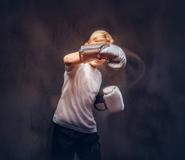 Schulbuben-Boxer mit blonden Haaren und weißem T-Shirt mit Boxhandschuhen zeigt einen Boxhaken. isoliert auf dunklem, strukturiertem Hintergrund. - Foto, Bild