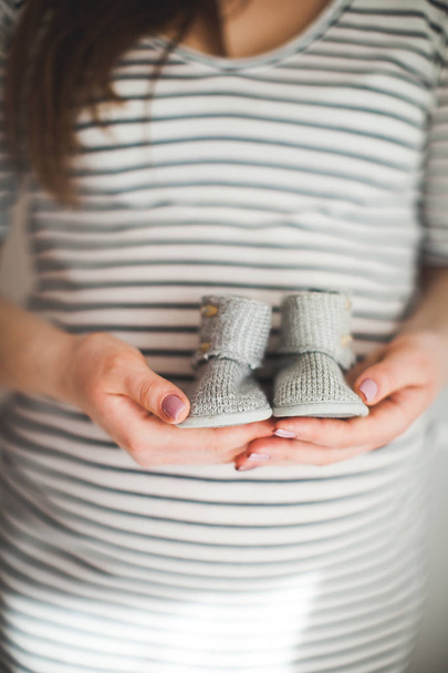 手は、小さな靴のペアを保持しています。灰色の背景に幼児の靴です。母親と赤ちゃんのテーマ。妊娠中の女性.  - 写真・画像