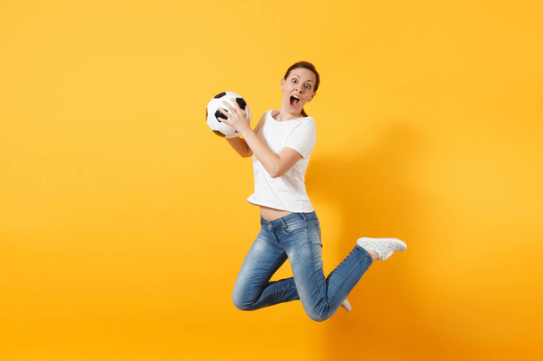 Nuori hauska ilmeikäs eurooppalainen naisjalkapallofani hyppää ilmassa, piristää tukitiimiä, pitäen jalkapalloa eristettynä keltaisella taustalla. Urheilu, pelata jalkapalloa, hurrata, fanit ihmiset elämäntapa käsite
 - Valokuva, kuva