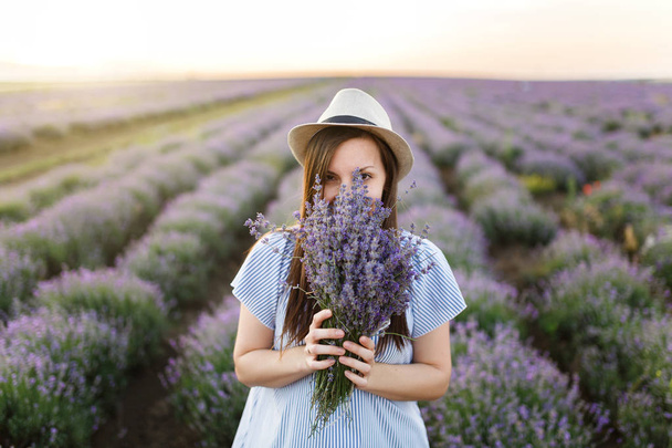 Young sensual beautiful woman in blue dress, hat on purple lavender flower blossom meadow field outdoors on summer nature background. Tender female near flowering bush with bouquet. Lifestyle concept - Fotografie, Obrázek
