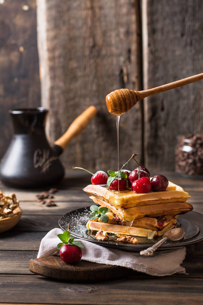 Freshly made belgian waffles with honey flows and powdered sugar. Cherries on top of waffles on wooden desk and napkin on wooden background. Coffee beans in glass jar. Turkish coffee pot for breakfast - Photo, Image