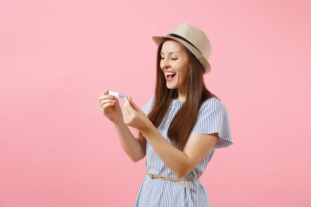Excited happy woman in blue dress, hat hold in hand, looking at pregnancy test isolated on pink background. Medical healthcare gynecological, pregnancy fertility maternity people concept. Copy space - Fotografie, Obrázek