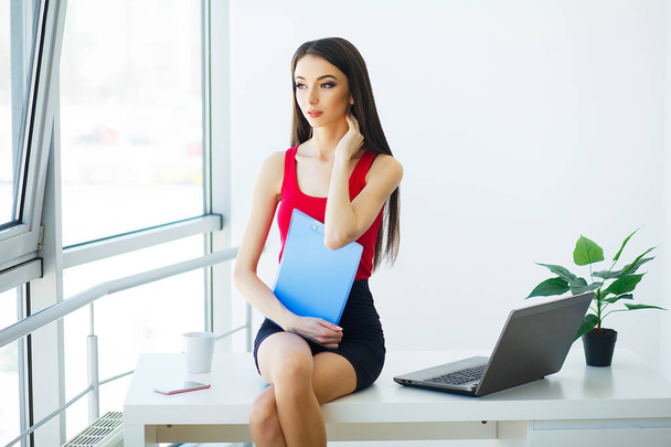 Бізнес. Портрет молодої жінки, що тримає блакитну папку. Одягнена в червону футболку та чорну спідницю. Ділова жінка сидить на столах у світлому сучасному офісі. Висока роздільна здатність
 - Фото, зображення