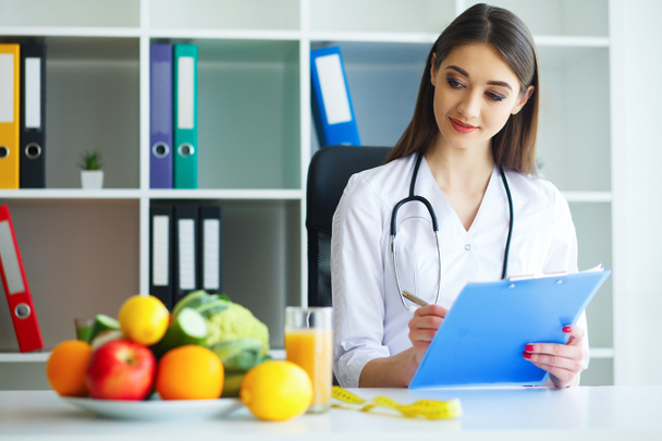 Health and Beauty. The Doctor Signs out a Diet Plan. The Nutritionist Holds in the Hands Fresh Fruits and Vegetables. Young Doctor with a Beautiful Smile at the Light Office. High Resolution - Photo, Image