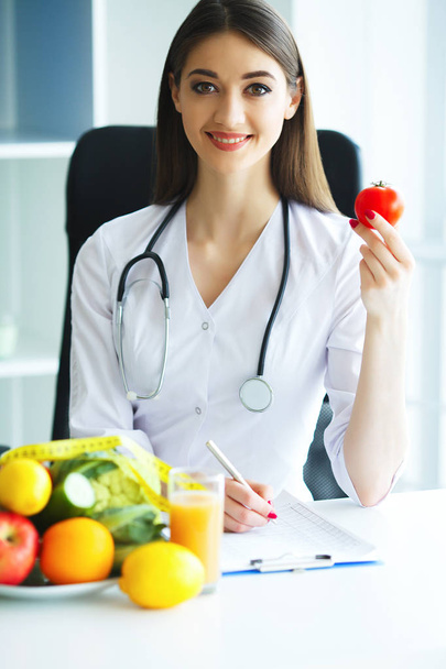 Zdrowia. Lekarz podpisuje się plan diety. Dietetyk trzyma w garści świeżych pomidorów. Owoce i warzywa. Młody lekarz z pięknym uśmiechem w urzędzie. O wysokiej rozdzielczości - Zdjęcie, obraz