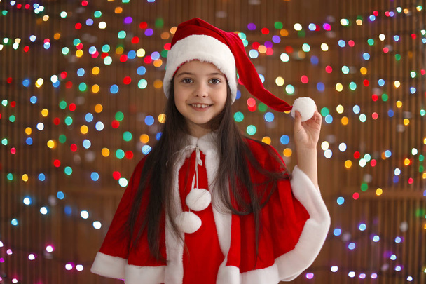 Cute little child in Santa hat on blurred lights background. Christmas celebration - Fotografie, Obrázek