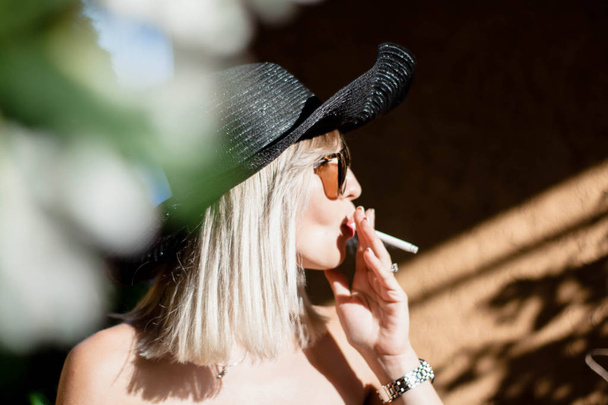 Beautyful elegante jovem loira mulher fumar cigarros ao ar livre em han e óculos de sol
 - Foto, Imagem