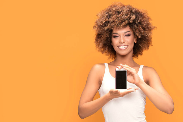 Photo of a smiling beautiful african woman with afro hairstyle, holding mobile phone with blank screen, standing over yellow background. - Photo, Image