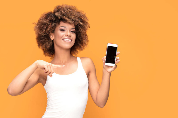 Photo of a smiling beautiful african woman with afro hairstyle, holding mobile phone and showing on blank screen, standing over yellow background. - Fotografie, Obrázek