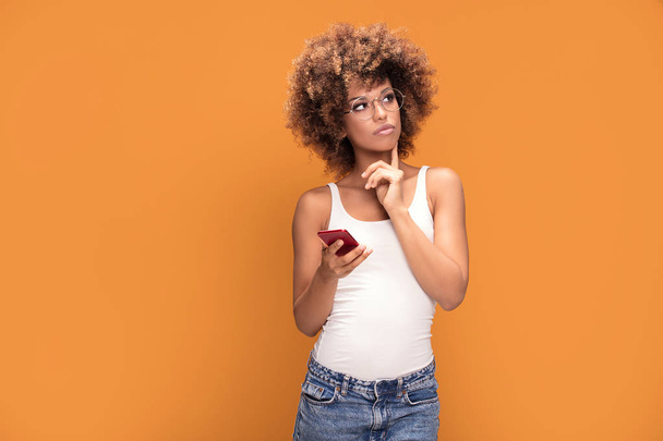 Photo of an african woman with afro hairstyle holding mobile phone, wearing fashionable eyeglasses and standing over yellow background. - Foto, Imagen