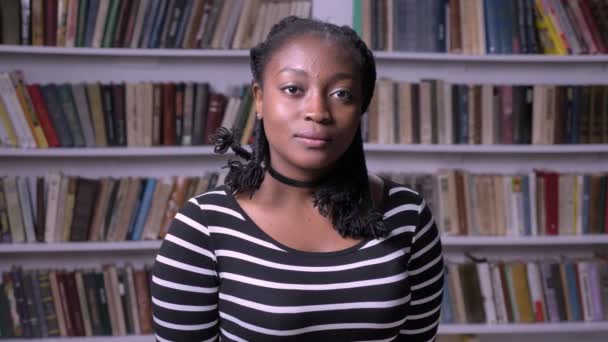 Young handsome african american woman standing in library and looking at camera, serious and thoughtful, bookshelves background - Footage, Video