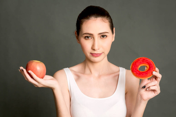 女性を保持していると灰色の背景に赤いリンゴとカロリー爆弾ドーナツの間で選択を行います。健康的な食事とジャンク フード コンセプト - 写真・画像