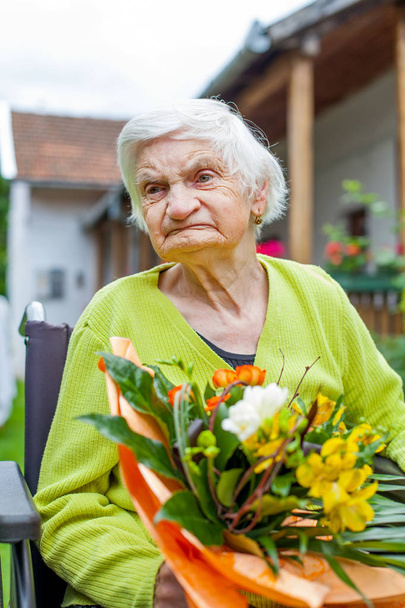 Disabled elderly woman sitting in wheelchair, receiving a flower bouquet for her birthday - Foto, Imagen