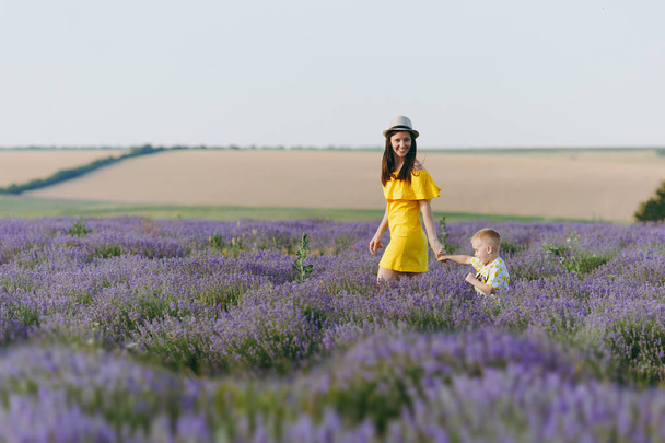 Young woman in yellow dress walk on purple lavender flower meadow field background, rest, have fun, play with little cute child baby boy. Mother, small kid son. Family day, parents, children concept - Photo, image
