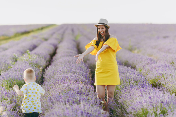 Young woman in yellow dress walk on purple lavender flower meadow field background, catch have fun, play with little cute child baby boy. Mother, small kid son. Family day, parents, children concept - Photo, Image