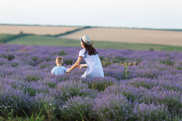 Young woman in blue dress walk on purple lavender flower meadow field background, catch up, have fun, play with little cute child baby boy. Mother, small kid son. Family day parents, children concept - Photo, Image