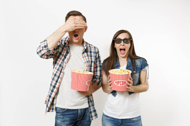 Young scared couple, woman and man in 3d glasses watching movie film on date holding buckets of popcorn covering faces with palm and screaming isolated on white background. Emotions in cinema concept - Φωτογραφία, εικόνα