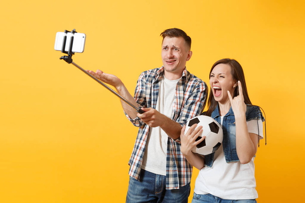 Jeune homme de couple femme, fans de football faisant selfie sur téléphone mobile avec bâton égoïste monopode, remonter le moral de l'équipe de soutien, ballon de football isolé sur fond jaune. Sport famille loisirs style de vie concept
 - Photo, image