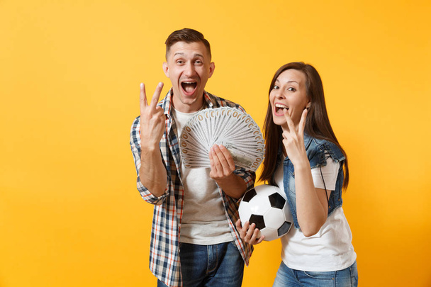 Young win couple, woman man, football fans holding bundle of dollars, cash money, soccer ball, cheer up support team isolated on yellow background. Sport bet excitement ardor family lifestyle concept - Photo, Image