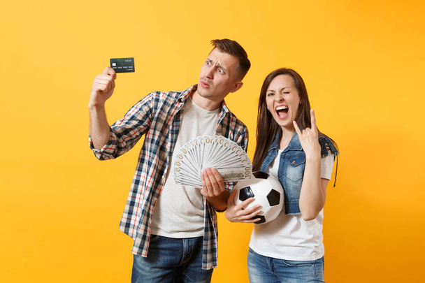 Young win couple, woman man, football fans holding bundle of dollars money, credit card, soccer ball, cheer up support team isolated on yellow background. Sport bet, ardor family lifestyle concept - Photo, Image