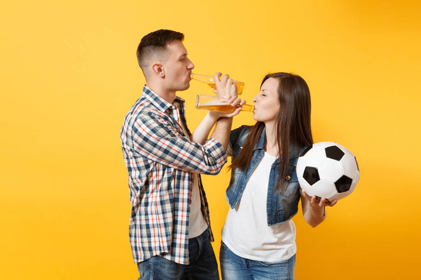 Joven pareja alegre simpatizante feliz, hombre mujer, aficionados al fútbol animar equipo de apoyo, sosteniendo botella de cerveza, pelota de fútbol aislado en el fondo amarillo. Deporte, ocio familiar, concepto de estilo de vida
 - Foto, Imagen