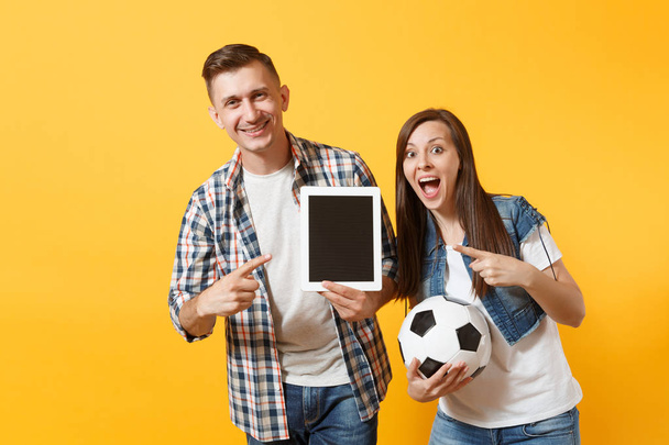 Νέοι διασκέδαση υποστηρικτής ζευγάρι, άνδρας γυναίκα, ποδόσφαιρο οπαδούς φτιάξει ομάδα υποστήριξης, κρατώντας κενή μαύρη μπάλα ποδοσφαίρου κενή οθόνη που απομονώνονται σε κίτρινο φόντο υπολογιστή tablet pc. Αθλητισμός, οικογενειακό τρόπο ζωής - Φωτογραφία, εικόνα
