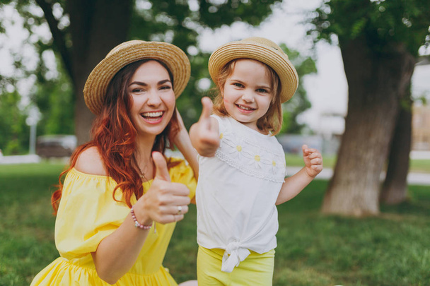 Smiling woman play on green grass lawn in park, have fun and showing thumbs up with little cute child baby girl. Mother, little kid daughter. Mother's Day, love family, parenthood, childhood concept - 写真・画像