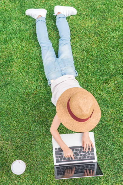 top view of child in hat lying on grass and using laptop - Φωτογραφία, εικόνα