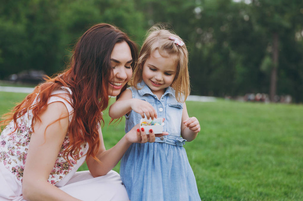 Mulher sorridente em vestido leve e pequena criança bonito bebê menina jogar, segurando caixa com papel confete no parque verde. Mãe, filhinha. Dia das Mães, família do amor, paternidade, conceito de infância
 - Foto, Imagem