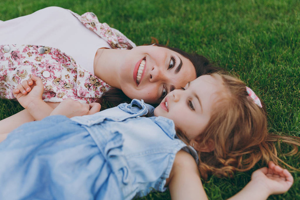 Smiling tender woman in light dress and little pretty child baby girl lie on green grass in park rest, have fun. Mother, little kid daughter. Mother's Day, love family, parenthood, childhood concept - Photo, Image