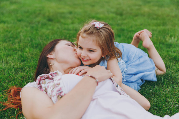 Tender woman in light dress kissing little cute child baby girl lie on green grass in park rest and have fun. Mother, little kid daughter. Mother's Day, love family, parenthood, childhood concept - Foto, Imagen