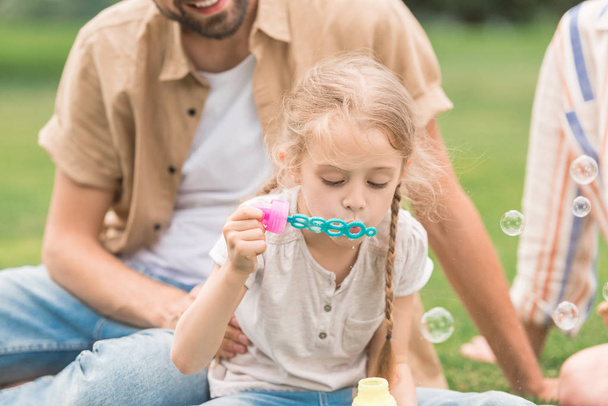 cropped shot of parents looking at cute little daughter blowing soap bubbles in park - Foto, Bild