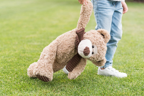 cropped shot of child holding teddy bear while standing on green lawn - Фото, изображение
