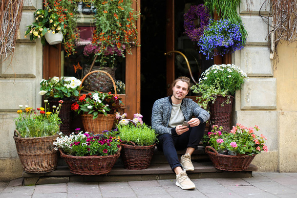Młody mężczyzna, trzymając smartfon i siedzi w kwiaciarni w pobliżu bukiety w kosze. - Zdjęcie, obraz