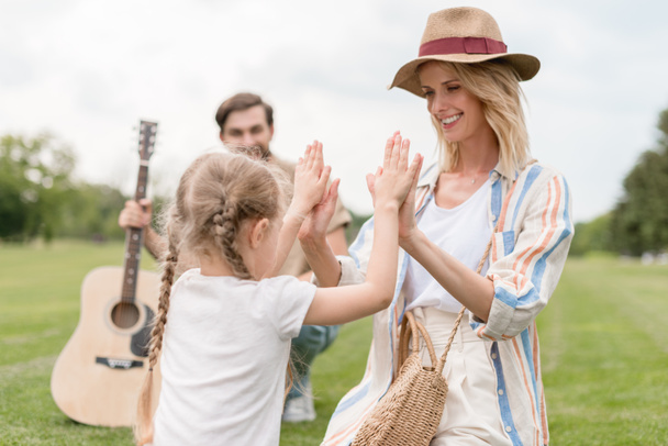 happy mother and daughter playing together while father holding acoustic guitar behind in park  - Foto, Imagem