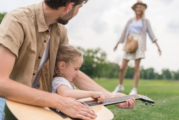 recortado disparo de padre e hija tocando la guitarra mientras madre de pie detrás en el prado
  - Foto, Imagen