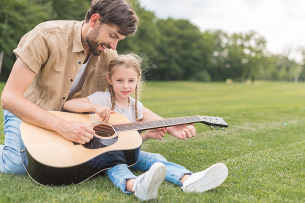 happy father and daughter playing acoustic guitar in park - Foto, afbeelding