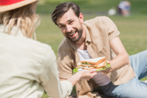 young couple holding sandwiches while spending time together at picnic in park - Φωτογραφία, εικόνα