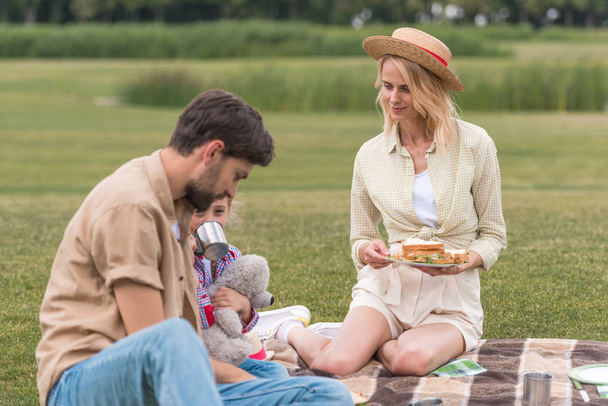 happy family with one child eating sandwiches while sitting on plaid at picnic - Photo, image