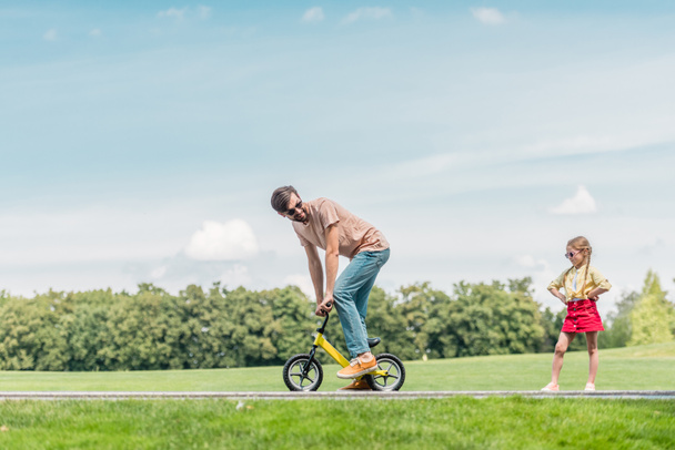 father riding small bicycle and looking at little daughter standing with hands on waist - Photo, image